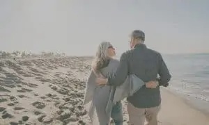 retired couple walking on the beach with arms wrapped around each other